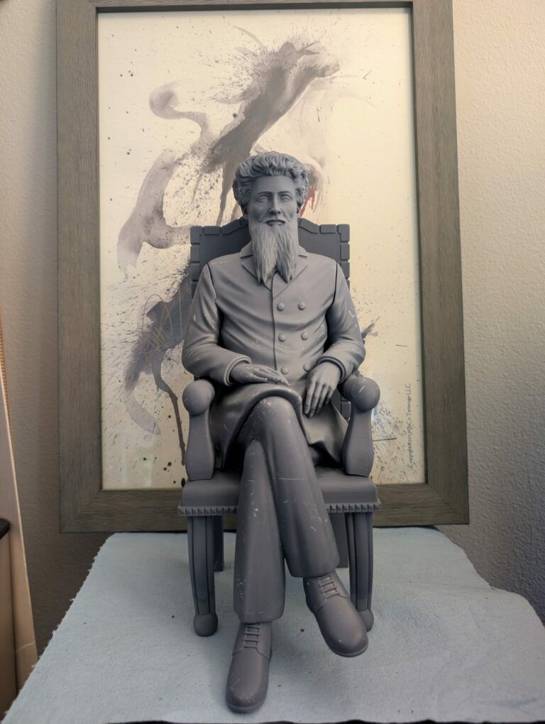 Grey statue of Emeterio Betances. He is a man with curly hair and a long beard. He sits in a large chair and crosses his legs.