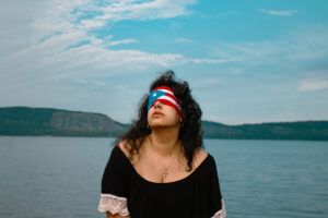 Amanda Raquel Dorval wears a Puerto Rican flag as a blindfold over her eyes. She is wearing a cross on a chain around her neck and a black dress with lace trim at the sleeve. Behind her there is expansive body of water and green trees. The sky is blue and filled with white clouds.