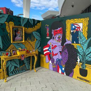 An installation view of a painting that is made up of two walls meeting like a room corner. The left wall is painted with vibrant green and yellow leaves. On the wall is a framed painting of a Taino person facing right. Underneath, there is a table populated by a fruit basket, picture frames, potted plants, and a small Puerto Rican flag. On the right wall, there is a painting of a Taino person braiding a black woman's hair. They are adorned by Puerto Rican flags.