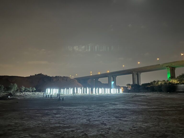 100 silver stainless steel columned light structures each standing 3 meters tall near a river at night.