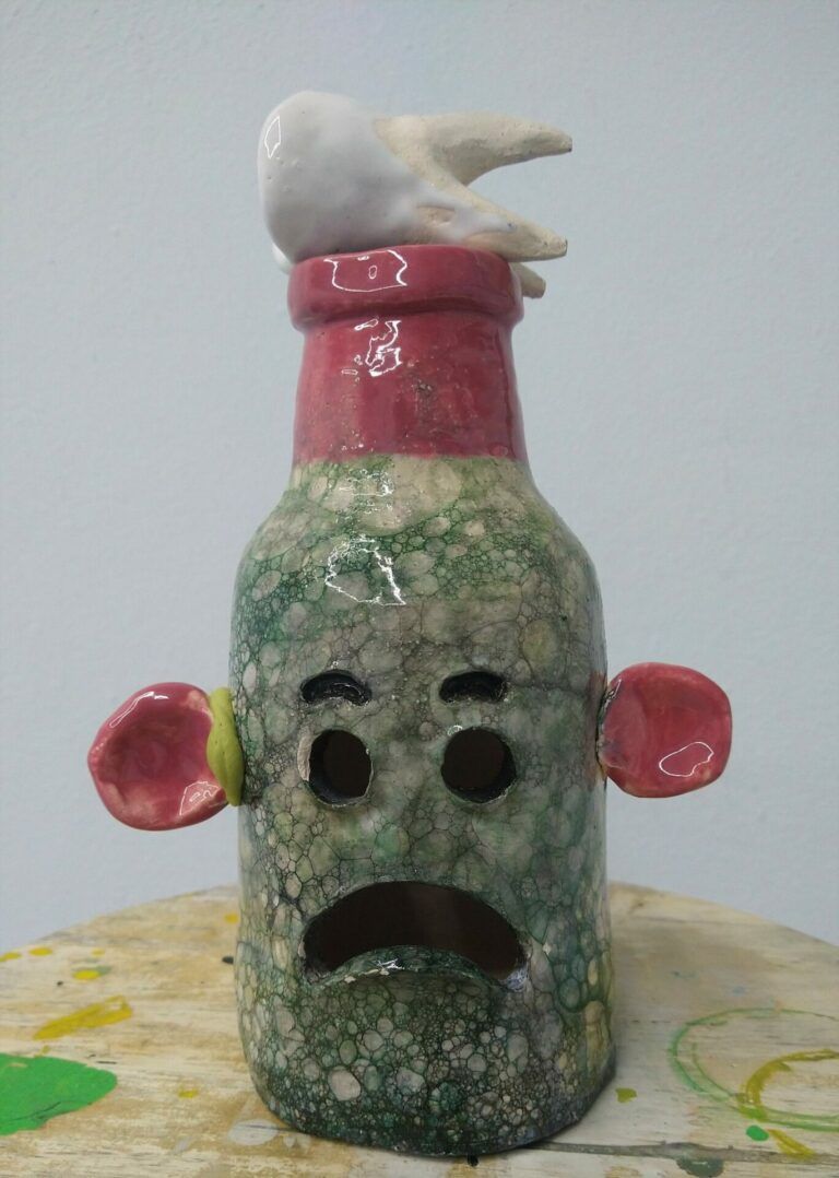 A bottle-shaped ceramic with pink ears and a cartoony frowny face. A big tooth sits on the top of the bottle.
