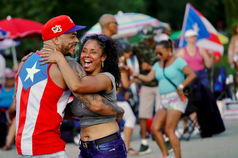 A man wearing a Puerto Rican flag shirt and a woman happily dance in the street. The background is full of joyful dancers.