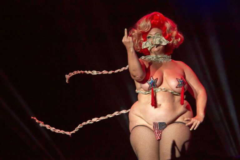 Drag queen dressed in red white and blue attire. Her wig is red and white. She wears nothing but an American flag covering her genitals and two star shaped pasties.