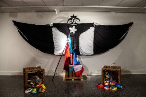 Black and white vejigante costume with arms outstretched, displaying a large cape under it's arms. On the floor there are spilled crates of vajigante masks.