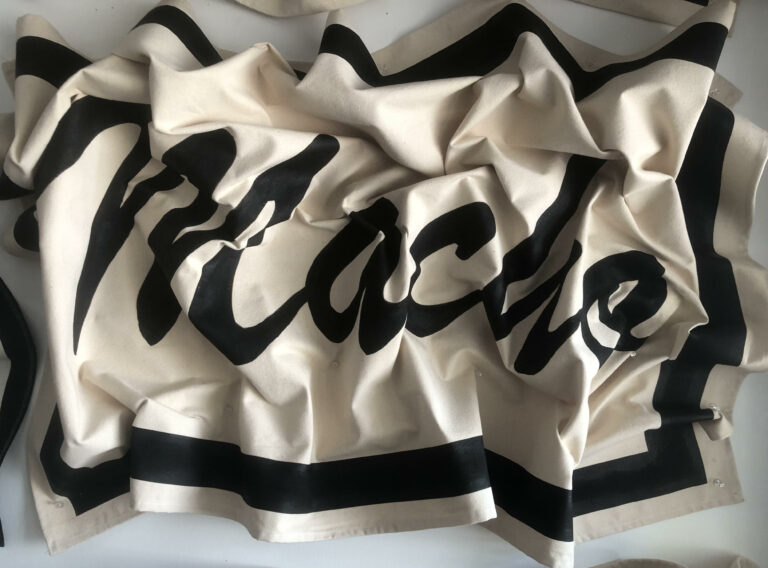 Black text "Macho" with a rectangular border on a cream colored canvas background. The canvas is crumpled but the text is legible.