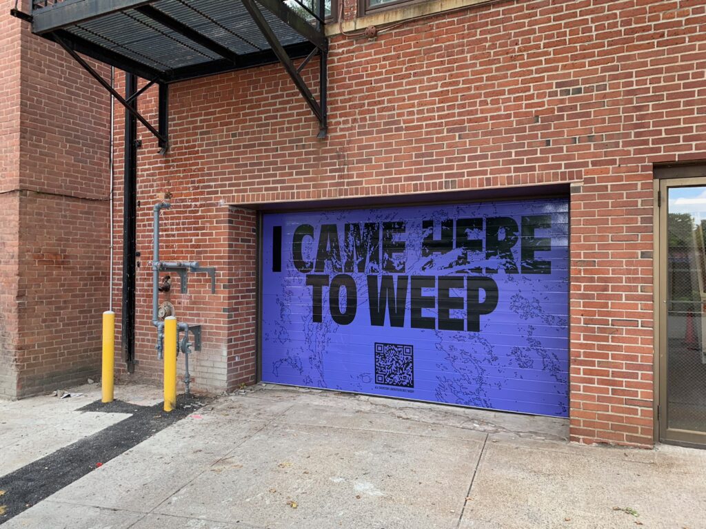 "I Came Here to Weep" printed in black text accompanied by a QR code on a garage door. The graphic includes a wave splashing effect.