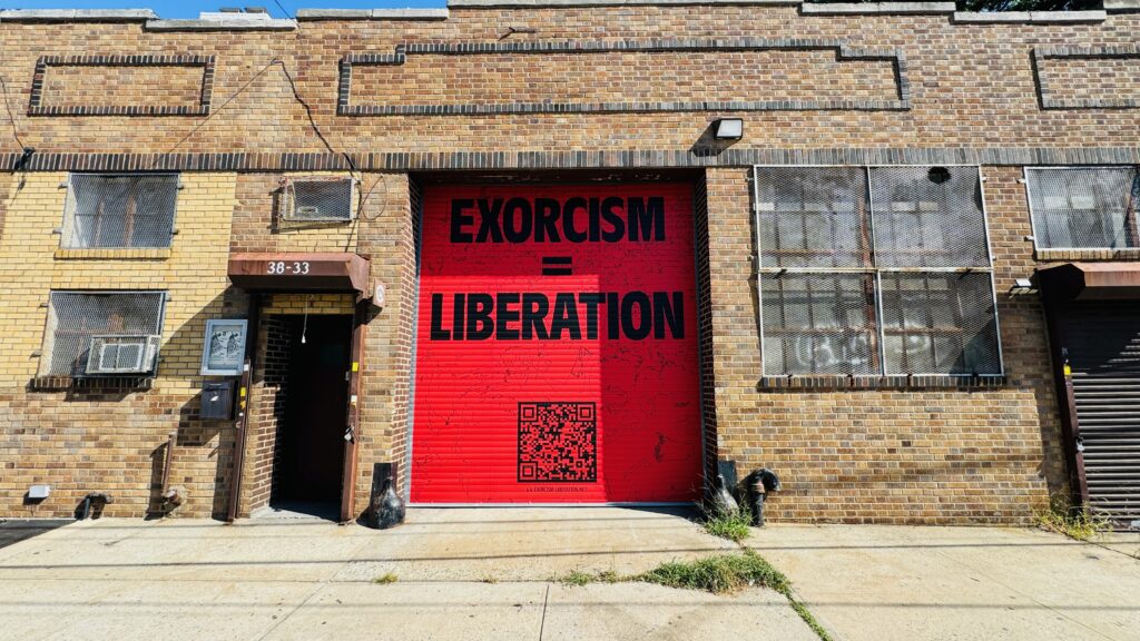 "Exorcism=Liberation" black text and QR code on red background. Painted onto a garage and has line-drawings of a crowd of people in the background.