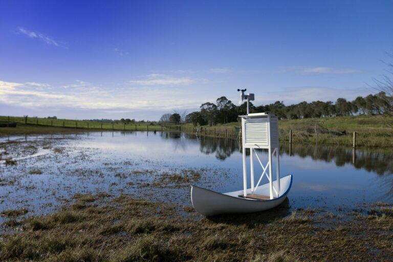 White canoe with a small weather station attached to the bench. The canoe sits in a shallow body of water.