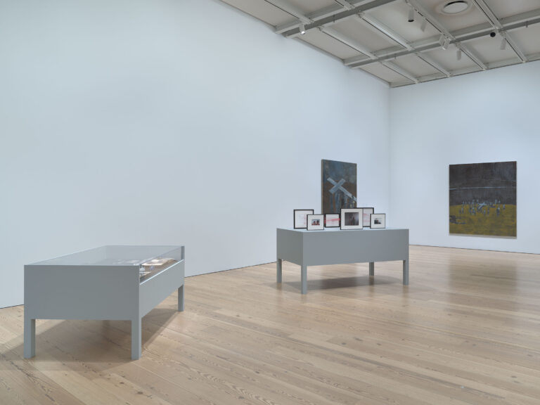 Image of a room at the Whitney Museum. There are two display cases and two paintings on the wall.