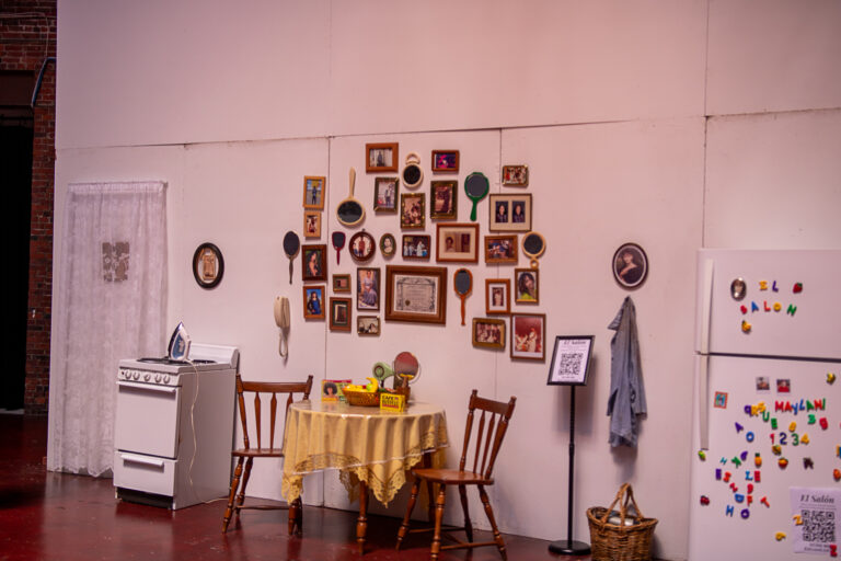 An installation of a fridge with magnets, an oven with an iron, a table with fruit and coffee, chairs, picture frames and mirrors.
