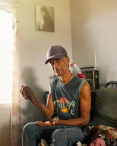 A descendant of the Tainos, Baracutey sits in his house holding up a wooden carved object with Taino symbols on it.