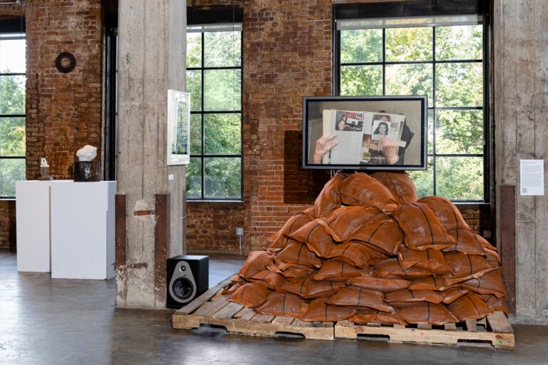 A image from the multimedia installation of "Un Dia Como Hoy." There are wood pallets holding up brown sandbags, a video screen right above it, and digital prints hung on the stone pillars in the large warehouse room.