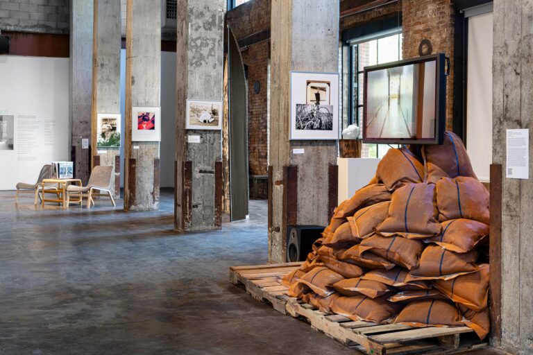 A image from the multimedia installation of "Un Dia Como Hoy." There are wood pallets holding up brown sandbags, a video screen right above it, and digital prints hung on the stone pillars in the large warehouse room.