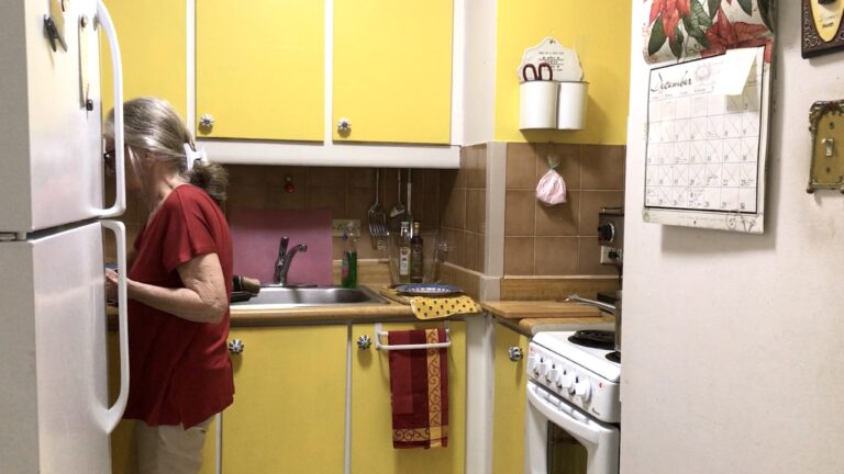 A kitchen with yellow cabinets, a white stove and fridge, and a calendar on the wall. Someone stands in the kitchen face obscured by the fridge, busy at the counter.