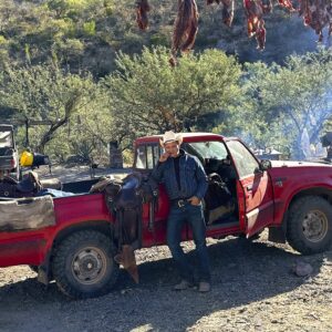 A photo of artist Werner Segarra leaning against a pickup truck. His hand is propped up next to his face and he is looking at something off camera with a neutral expression.