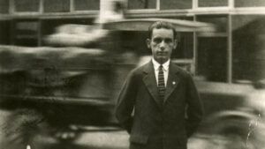 A young person in a suit stands in front of a street with a car speeding past.