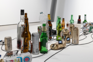 Crushed cans and bottles are lined up against a white background and intertwined with black cables.