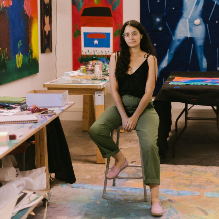 A headshot of artist Yanira Vissepo seated in her studio.She wears glasses and smiles lightly at the camera.