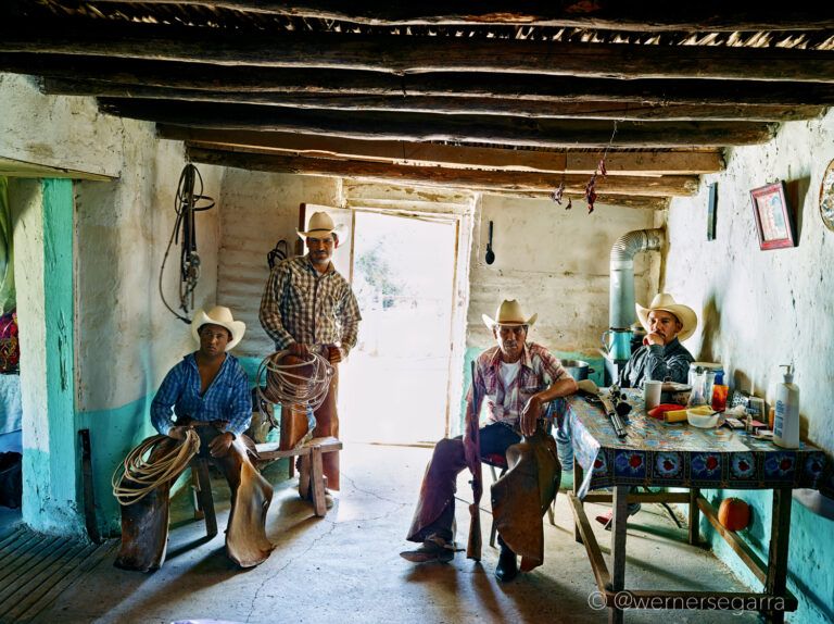Four men sit and stand around a living space. They all wear their vaquero hats and chaps, lasso included. They sternly look at the camera.