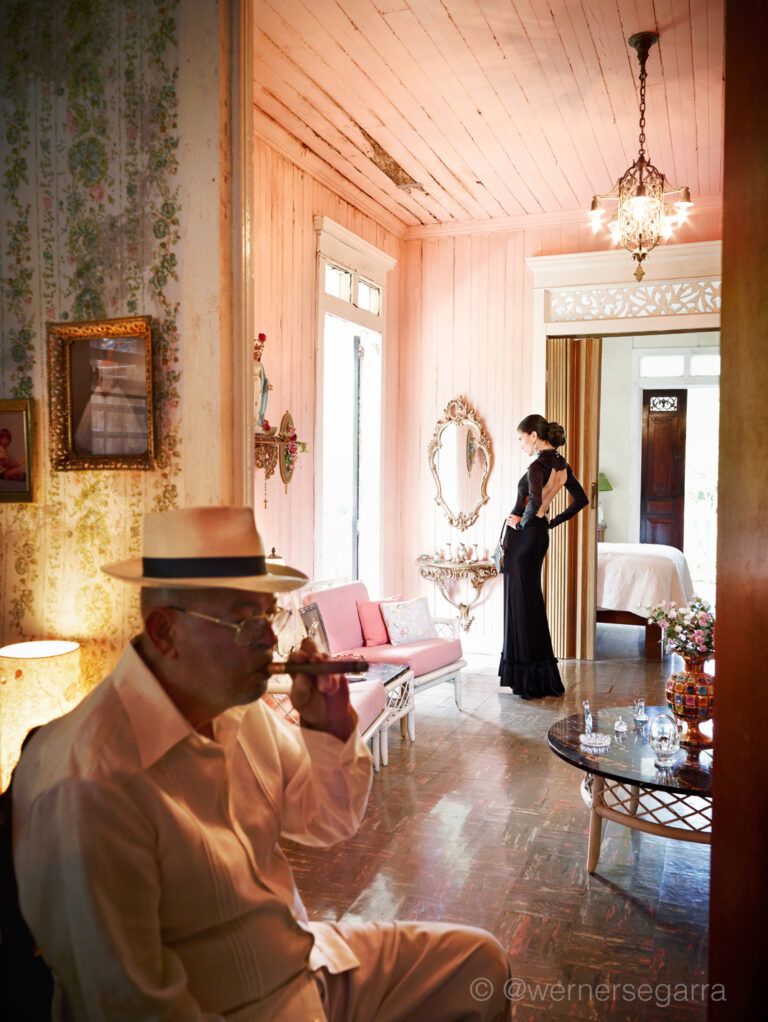 A woman stands facing a window in the background. She wears a backless gown. In the foreground, an older man smokes a cigar. They are both in a nice looking home.