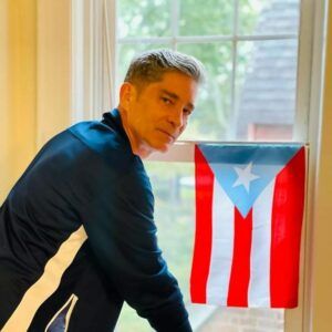 A photo of artist Ángel Vázquez standing by a small Puerto Rican flag. He turns and looks to the camera with a neutral expression.