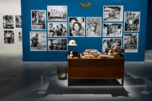 The blue and white walls of the Museum of the Old Colony are covered in black and white photographs of people, statues, and monuments. In the center of the room there is a full desk covered in objects. The desk is accompanied by a trash bin. It is roped off so that museum-goers do not interact with it.