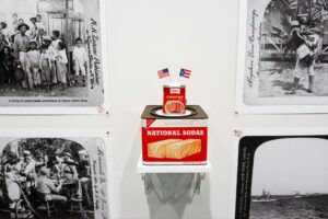 Inside the Museum of the Old Colony, the installation walls is white. There are old black and white photos with text in script on the sides. In the center, there is a can of "National Sodas" and a small can of "Corned Beef" sitting on top with the American and Puerto Rican flags sticking out.