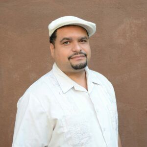 A photo of artist Dennis Flores. He wears a white hat and shirt and smiles at the camera.