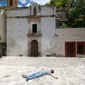 Artist Jason Mena in front of old buildings. Jason is lying face up on the street with arms out.