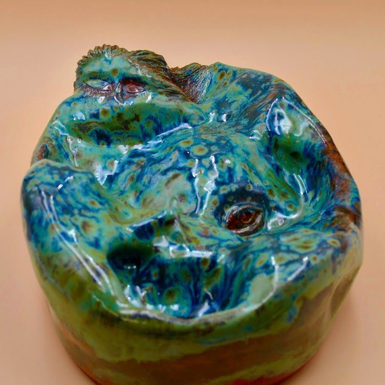 A rounded green-blue glazed ceramic with faces carved into the clay.