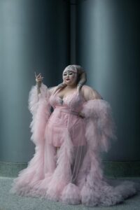 "This digital photograph, taken in 2022 at a Star Wars convention in Anaheim, California, features a plus-size fan cosplaying as a Twi'lek." - Rhynna Santos. The fan is wearing a pink frilly floor-length nightgown. Her lekku (head appendages) are a matching pink color.