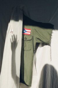 A shadow reaches towards a shiny dog tag hanging from a disembodied uniform with a Puerto Rican flag patch.