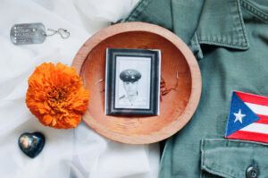 A wooden bowl with an image of an officer inside, a marigold flower, a heart shaped stone, a dog tag, and a uniform laid out on white fabric.