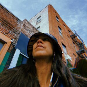 Artist Vanessa Rodriguez stands in front of brick buildings and looks upward.