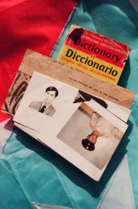 An English-Spanish dictionary, a print that reads "We see through the eyes of our ancestors," and a few photos of a man, one in uniform, one without, all resting on a Puerto Rican Flag.