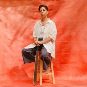 Artist Cándida González photographed by Diana Albrecht. They are sitting on a stool in front of an orange backdrop.