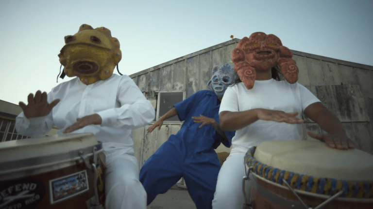 Two drummers wearing Taino style masks play for a mask-wearing dancer in blue.