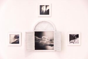 Four black-and-white photographs on a light pink background. The centerpiece is a light pink window with shutters open that contains one of four photos. The photos show a child and the sky.