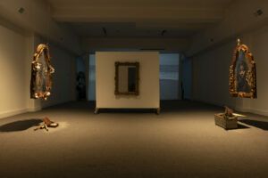 Two portraits facing each other in a dim gallery. Under each portrait there is a wooden box with small objects inside. Between the opposing frames, facing the viewer there is a mirror.