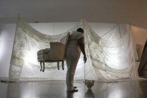 A still from a performance installation. A white room with white curtains and white furniture is being rearranged by someone wearing a white jumpsuit.