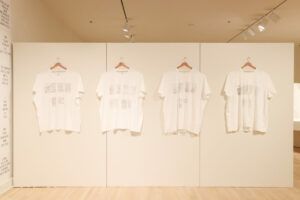 White T-shirts lined up on hangers on the wall. Literature pages are printed onto the shirts in black.