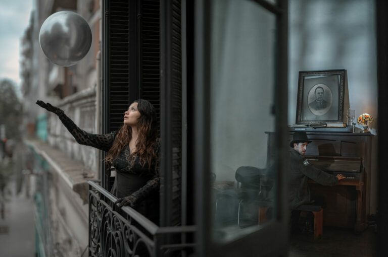 A woman wearing all black reaches out for a silver ball floating outside her balcony. A man inside plays the piano.
