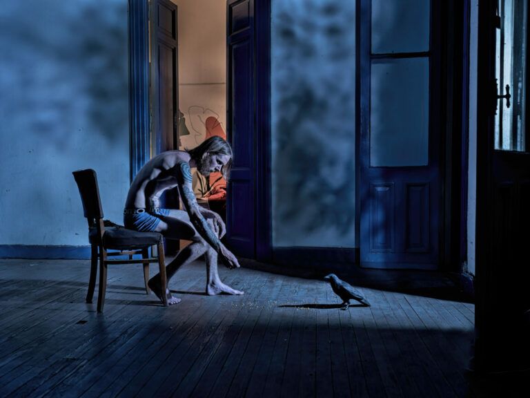 A person reaching out to a bird in a blue room. In the distance, an open room is lit orange,