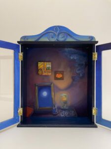 A blue wooden cabinet with paintings and keepsakes inside.