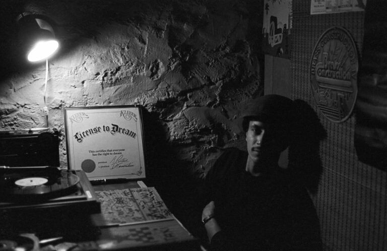 Man in a dark room with a record player and a framed certificate that says "License to Dream."