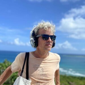 Artist heryk tomasini-suro standing in front of an overlook of the ocean. He wears headphones and glasses.