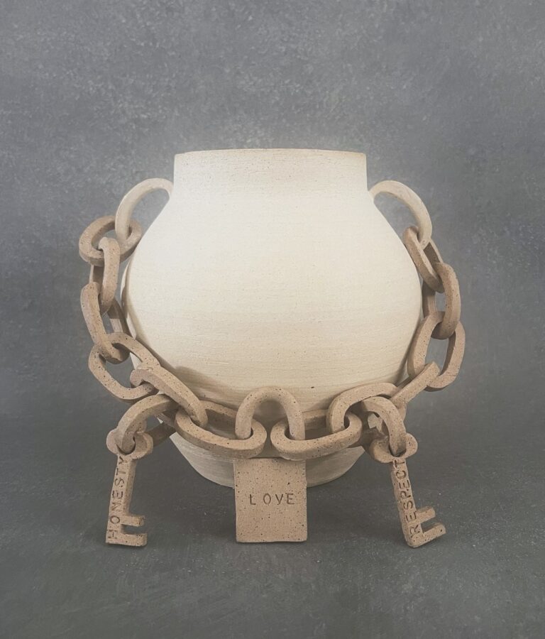 A stark off-white vase with rounded sides and two handles. Connecting the handles is a chain link with a lock form in the center that reads "Love." Two keys are attached to either side of the lock. The left reads " Honesty" and the right reads "Respect."