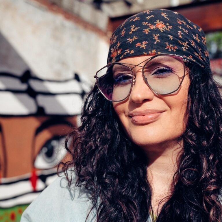 The artist Elizabeth Baez smiles at the camera. She wears a bandana and glasses. Her dark hair is big and wavy.