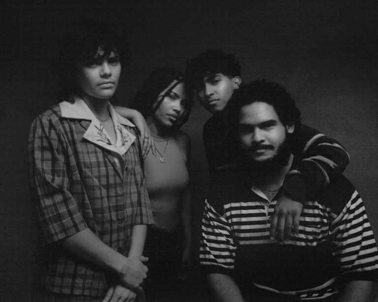 Black and white photo of the band Chuwi. Four people stand leaning on each other in heavy shadow.