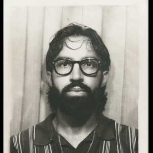 Black and white artist headshot. Daniel Delgado looks at the camera head on. He wears a striped shirt and dark glasses.